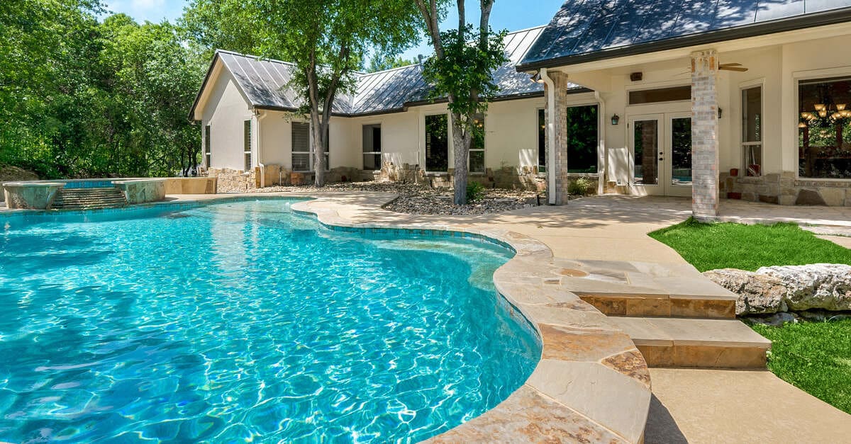 A backyard swimming pool on a sunny day. The pool has stone pavers and steps. A home and trees surround the area.