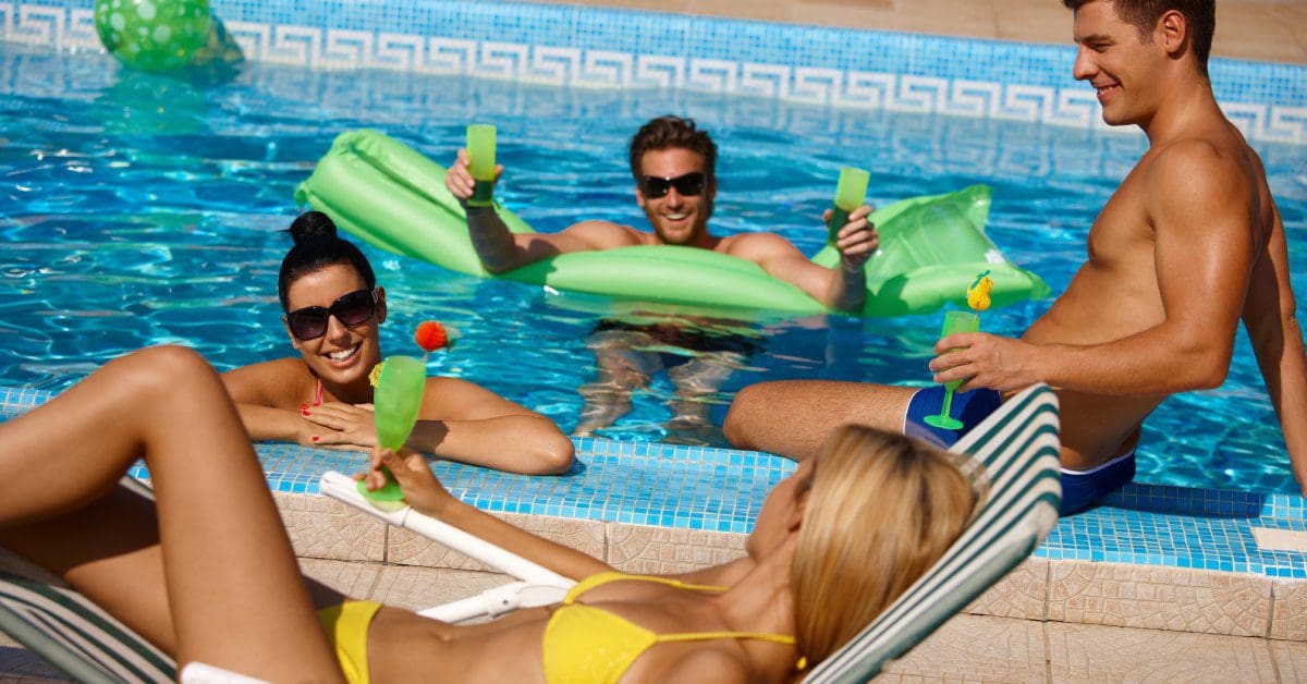 Four young adults in bathing suits lounging in and around a pool. All but one are holding drinks in green glasses.