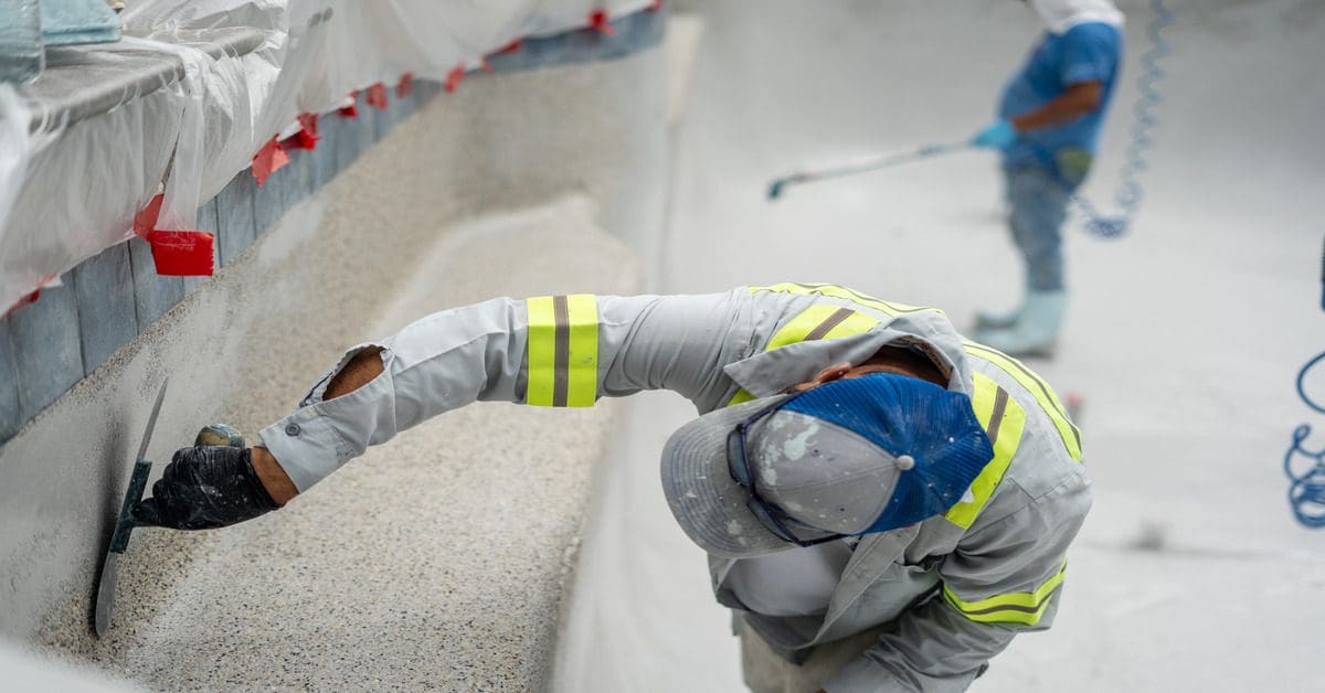 A close-up of a guy repairing a swimming pool by plastering the sides. They're wearing highly visible safety gear.