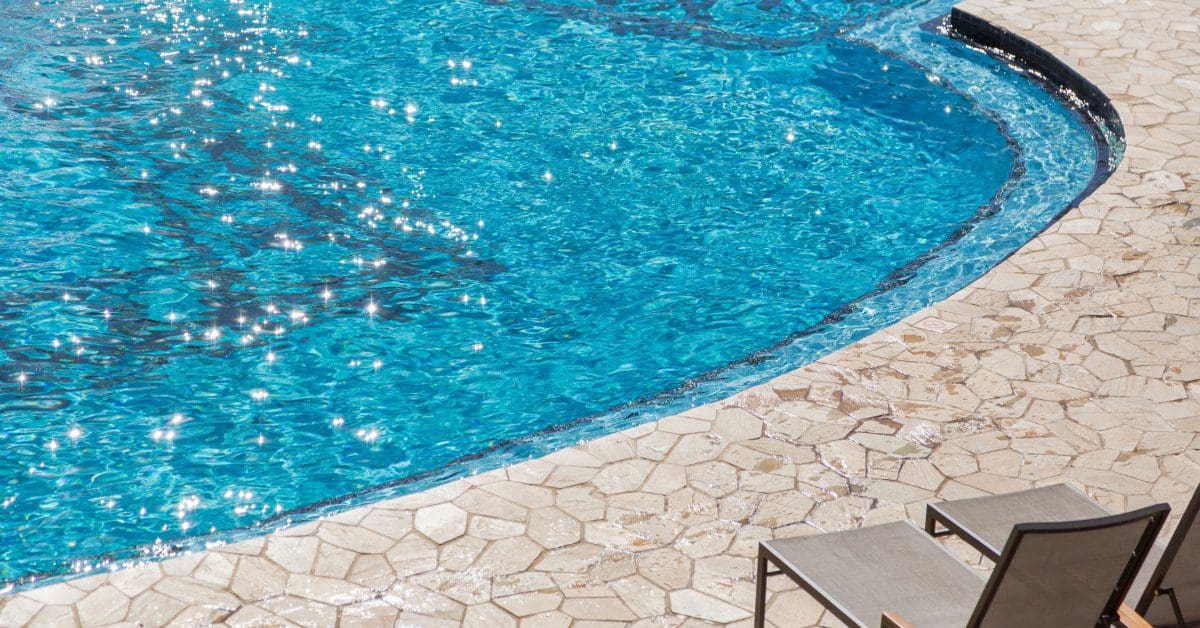 A close-up of the edge of a residential poolside. Two chairs are sitting on the stone walkway at the pool's edge.